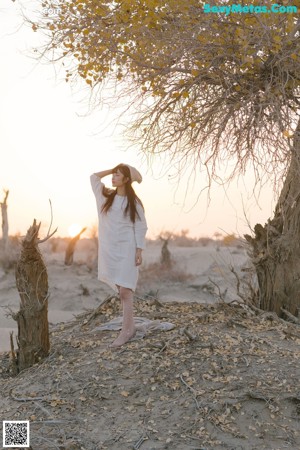A woman in a white dress and hat standing next to a tree.